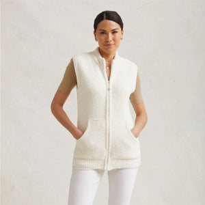 A woman stands against a light gray background wearing the KASHWERE - UNISEX VEST by KASHWERE, a sleeveless, zip-up cream-colored knit vest over a short-sleeve beige top. She has her hands in the cozy layering pockets and is dressed in white pants. Her dark hair is pulled back as she looks at the camera with a slight smile.