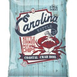 A small plate of kettle-cooked potato chips is shown in close-up, with a 5oz bag of Carolina Kettle - Crab Boil Chips by 1 In 6 Snacks in the background. Nearby, a metal container filled with chips and seashells captures the essence of a Carolina summer, celebrating southern tradition.