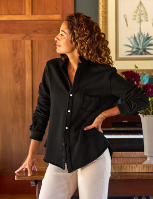 A person with curly hair smiles while standing next to a wooden table, wearing the EILEEN Relaxed Sweatshirt Button Up by FRANK & EILEEN, along with white pants. In the background are wooden panels, framed botanical artwork, and a piano adorned with flowers, creating a serene scene.