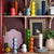 The kitchen shelf showcases ADDISON ROSS LONDON Grande Salt or Pepper Mills, available in vibrant hues of red, yellow, blue, green, and orange—each designed with a durable ceramic mechanism. The shelf is also adorned with plates, jars, a small potted plant, a painting, and pastel-colored decorative items set against a brown wall background.