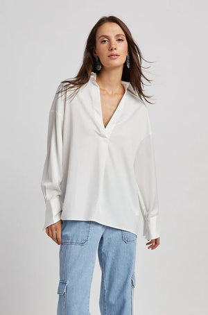 A person with long brown hair is standing with their back partially turned, wearing the ADROITE ATELIER - KAY POPOVER SATIN BLOUSE, an oversized white V-neck satin blouse from ADROIT ATELIER, paired with light blue jeans. The person is also wearing large, ornate earrings. The background is plain and light-colored.