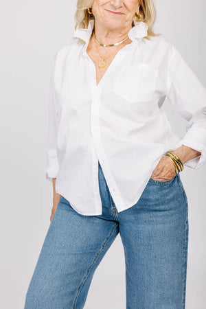 A woman is pictured from the shoulders down, wearing the versatile KMJ - QUARANTINA SHIRT, a white button-up with rolled-up sleeves, paired with blue jeans. She has her hands in her pockets and accessorizes with layered gold necklaces and bracelets. Her nails are manicured and painted white.