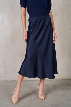 A person wearing a navy blue SATIN MIDI SKIRT from ROSE & CROWN paired with a matching navy blue top. The individual is also wearing beige pointed-toe heels. The background features a plain, light-colored wall with a concrete floor.