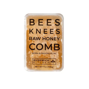 A rectangular tin container labeled "BUSHWICK KITCHEN - 'BEES KNEES' RAW HONEYCOMB" by Bushwick Kitchen. The off-white tin with black and orange text states "Born in Brooklyn, NY" and "Net Wt 7oz (198g)." One side is marked "100% Raw Honeycomb," containing pure, edible honey comb.