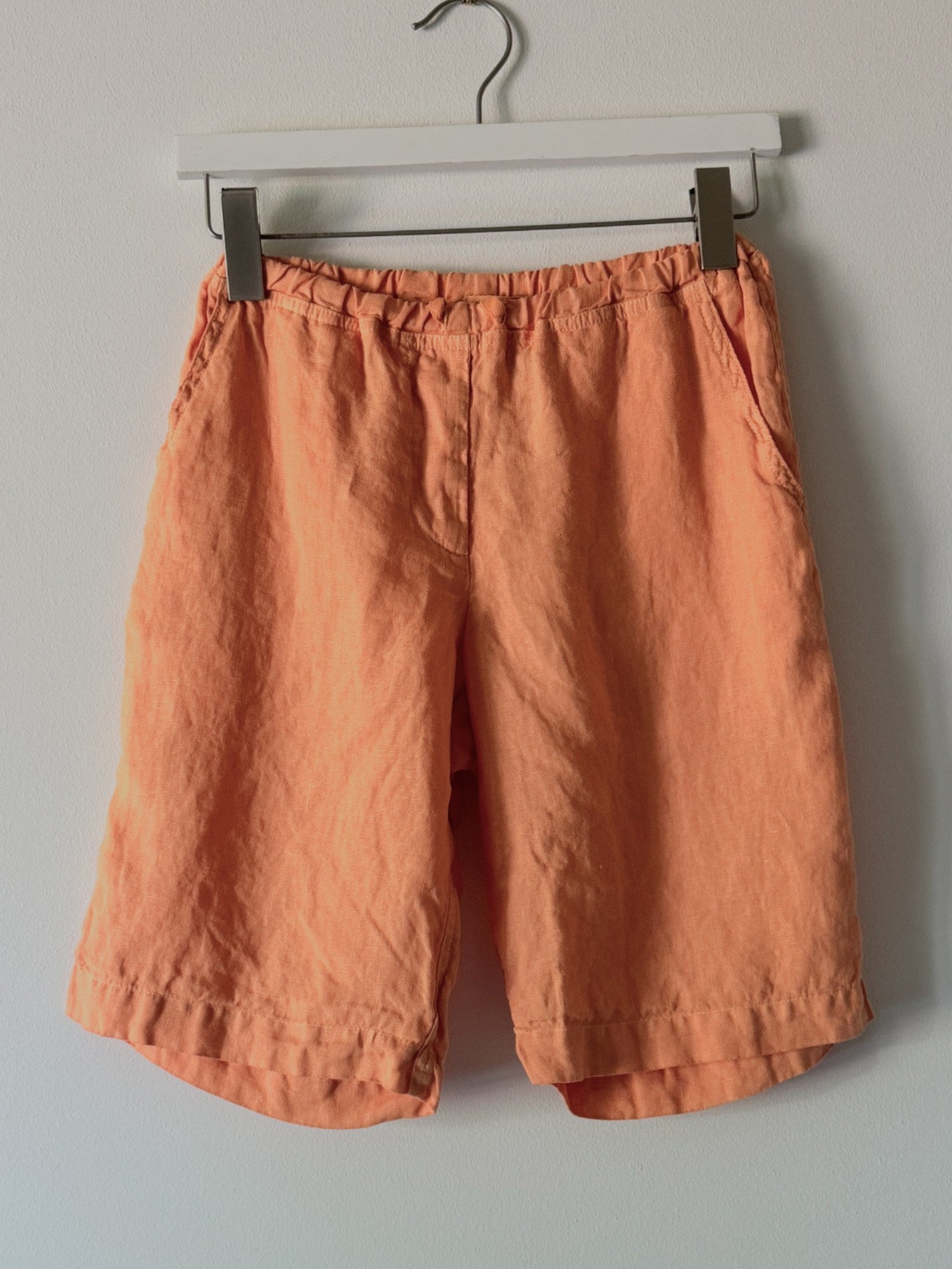 The CP SHADES - HAMPTON SHORT in orange linen is hanging on a white plastic hanger against a white wall. These drawstring shorts from CP SHADES feature an elastic waistband and side pockets, made from high-quality linen fabric.