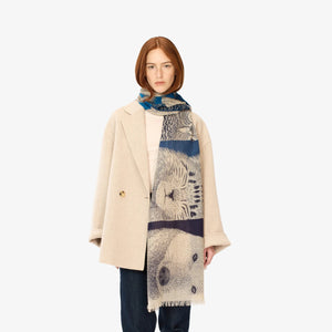 A person with shoulder-length red hair is wearing a light beige coat over a light-colored top, dark pants, and the INOUI EDITIONS - FRERES SCARF by INOUI, which features a detailed face design and is perfect for winter warmth. The background is plain white.