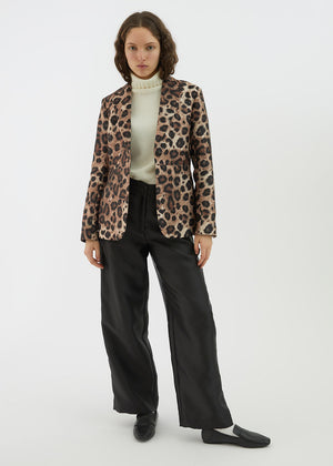 A person with wavy brown hair is wearing the ALYKI LABORATORIO LEOPARD PRINT JACKET over a white turtleneck sweater. They have their arms crossed and are standing against a plain white background.