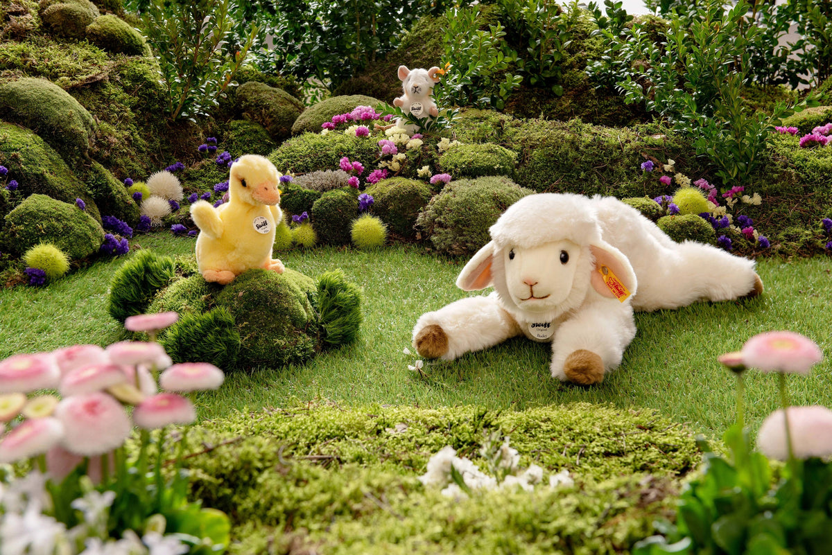 A plush lamb rests on a grassy patch with STEIFF&#39;s PILLA DUCKLING perched on a mossy mound. A small dog toy peeks from behind bushes, surrounded by vibrant flowers and greenery, creating an enchanting gift scene.