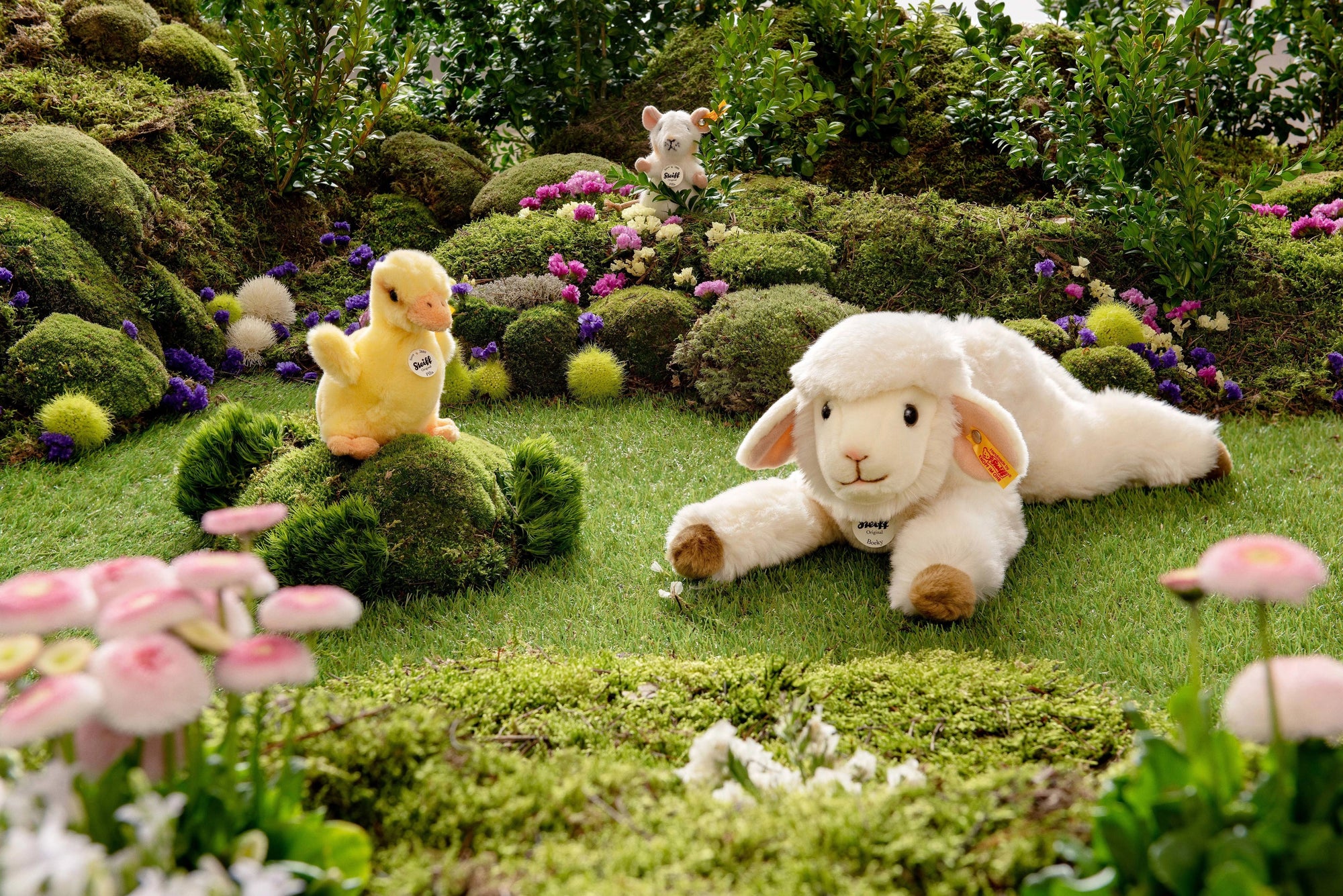 A plush lamb rests on a grassy patch with STEIFF's PILLA DUCKLING perched on a mossy mound. A small dog toy peeks from behind bushes, surrounded by vibrant flowers and greenery, creating an enchanting gift scene.