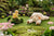 A plush lamb rests on a grassy patch with STEIFF's PILLA DUCKLING perched on a mossy mound. A small dog toy peeks from behind bushes, surrounded by vibrant flowers and greenery, creating an enchanting gift scene.