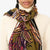 Close-up of a person wearing a cream turtleneck sweater and the INOUI EDITIONS - WESTERN SCARF from INOUI, featuring an abstract horse design. The washable wool scarf is predominantly olive green with accents of purple, red, and black. The person's face is partially visible with a calm expression.