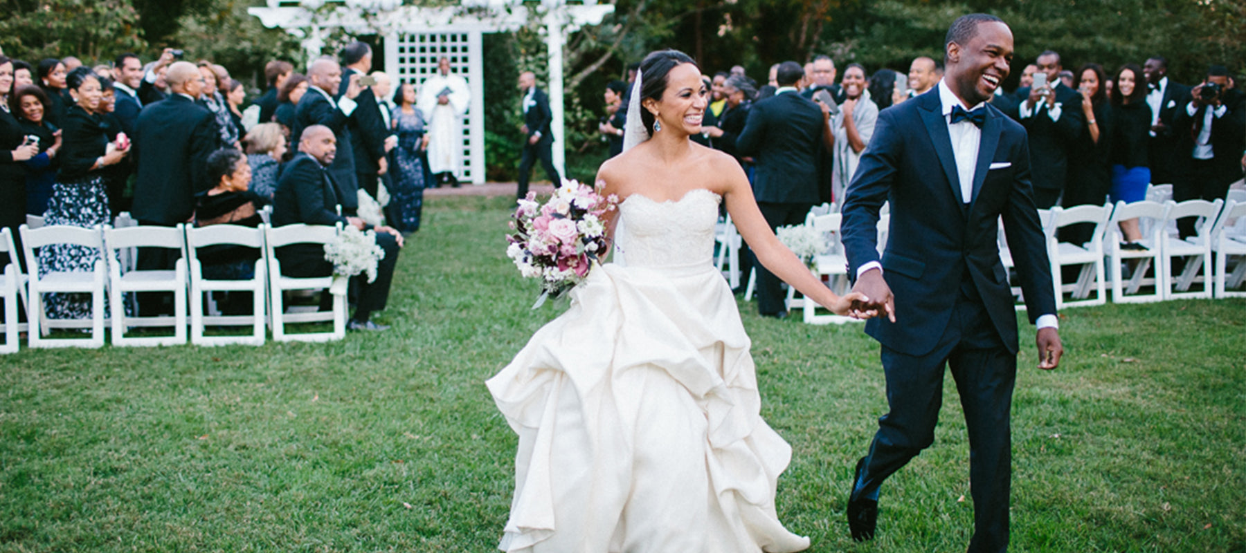A bride in a white gown and a groom in a black tuxedo walk hand in hand down an outdoor aisle, smiling, with guests seated and watching in the background.