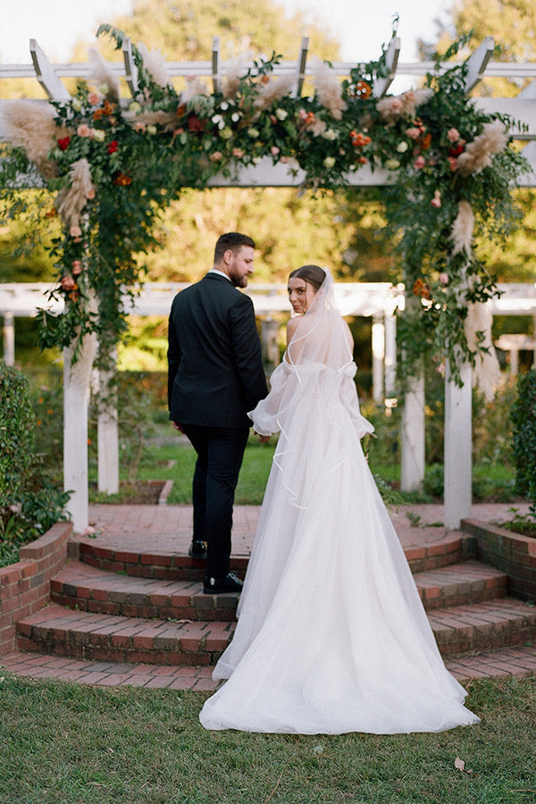 A bride in a white gown and veil holds hands with a groom in a black suit as they walk up brick steps under a floral archway in a garden setting. Both look back toward the camera.