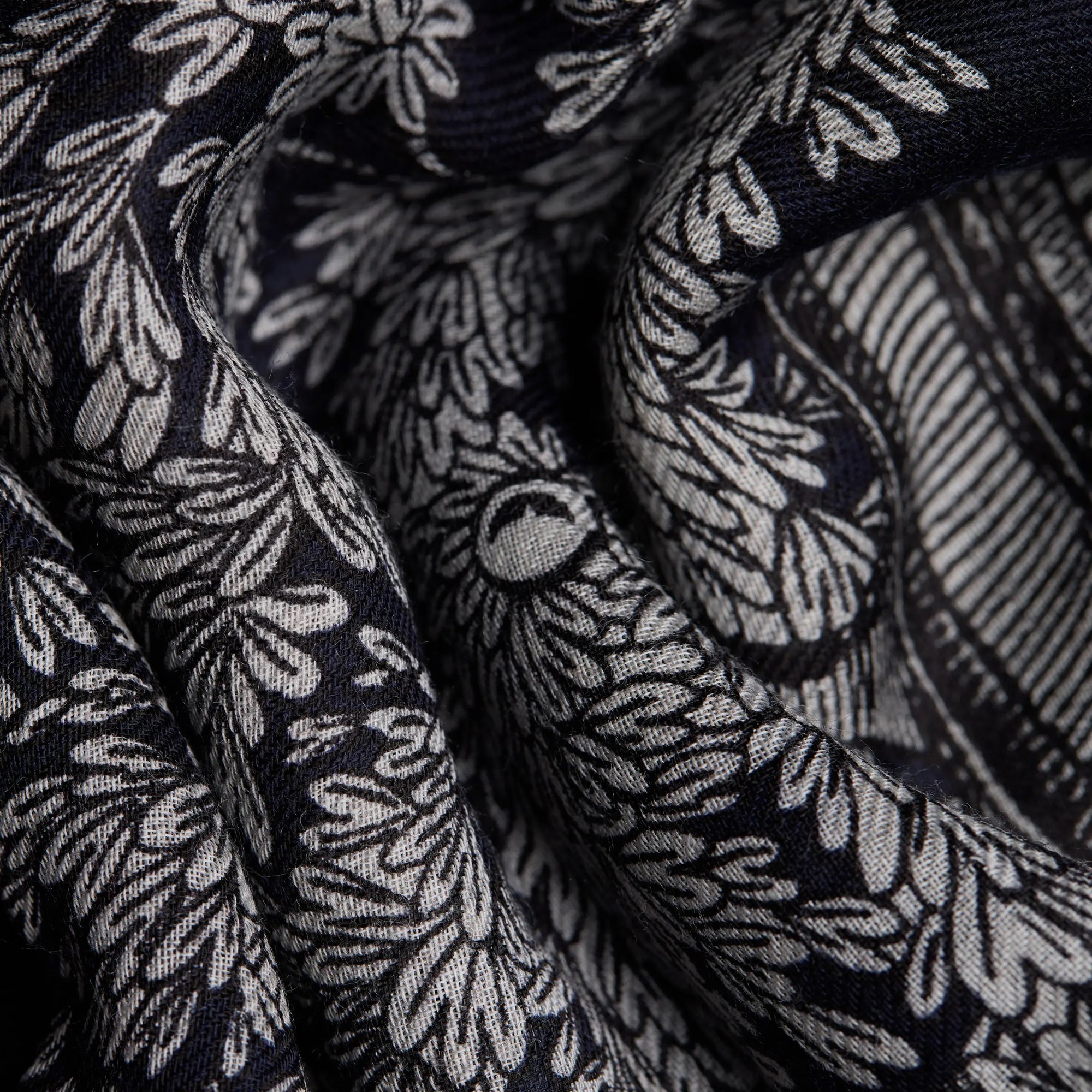 Close-up image of the INOUI EDITIONS - MIRAGE SCARF by INOUI, showcasing its intricate white floral and leafy patterns. The wool fabric is softly folded, creating gentle curves and shadows that highlight its unique design, making it an exquisite summer accessory.