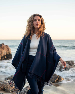 A woman with wavy brown hair drapes the MERSEA - PLAID POCKET WRAP, featuring fringe and dark stripes, over her black top and pants. This versatile travel wrap from MER SEA is ideal for any journey, providing both style and easy care as she stands against a plain light background, holding it around her shoulders.