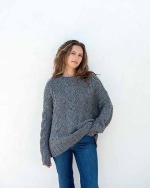 The image displays a detailed view of a person dressed in the MERSEA - TOPANGA CABLE SWEATER by MER SEA, showcasing its chunky knit cream-colored texture with large buttons on the sides, paired with blue jeans. The emphasis is on the sweater's intricate cable design and fine details, with the person's hand partially visible at their side.