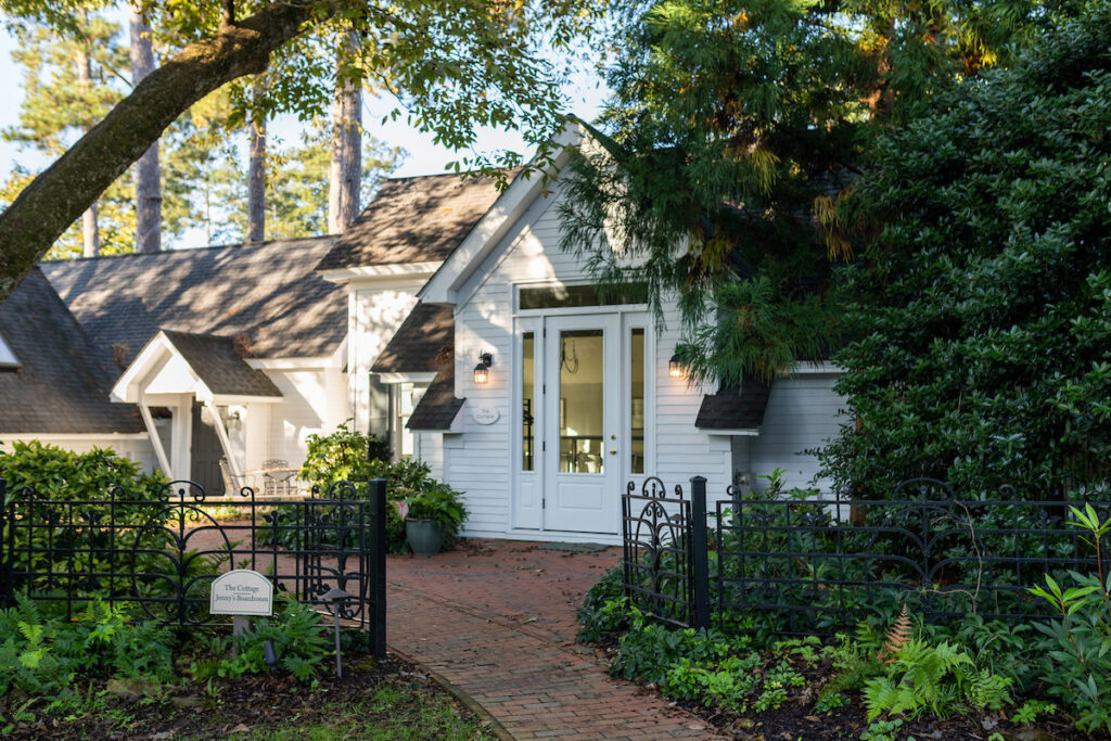 A charming white cottage with a steep roof, large windows, and a glass door sits behind a black wrought-iron fence and lush greenery, with a brick path leading to the entrance on a sunny day.