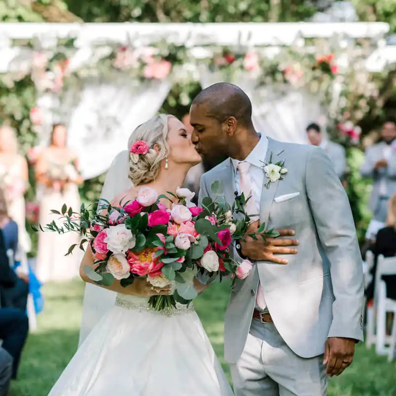 A bride and groom kiss outdoors at their wedding ceremony; the bride holds a large bouquet of pink and white flowers, and both are dressed in formal wedding attire, surrounded by greenery and floral decorations.