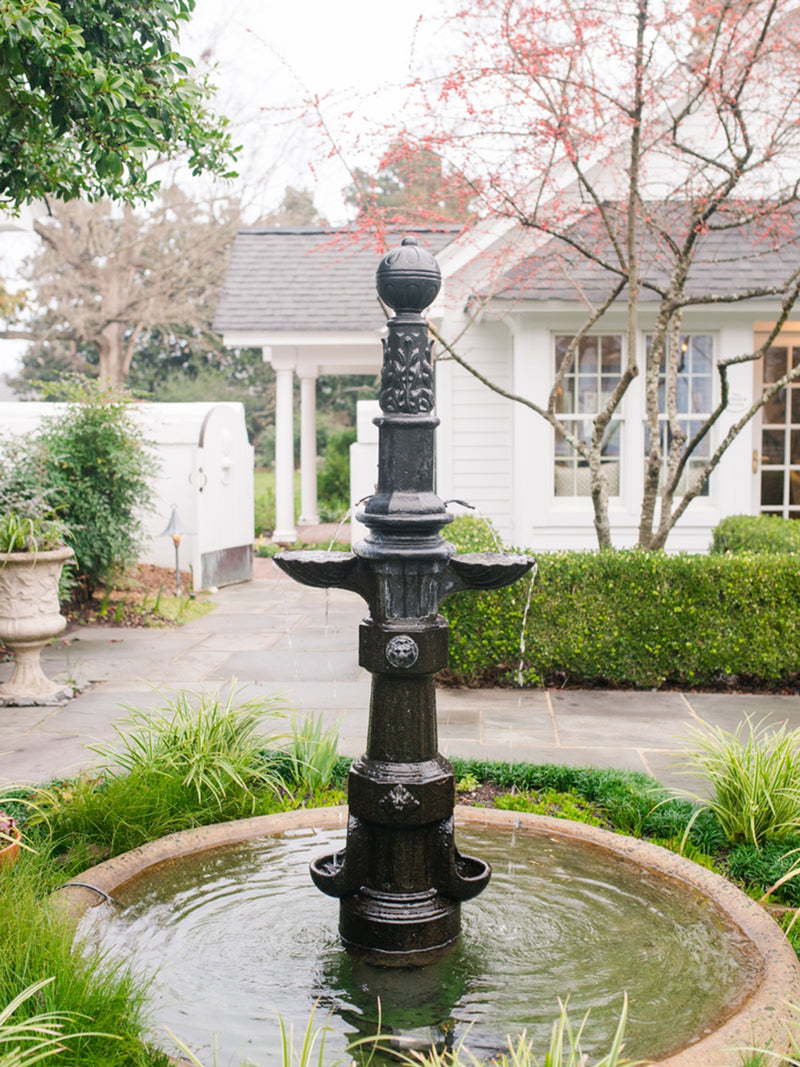 A tall, ornate black fountain stands in the center of a round pond surrounded by greenery, with a white house, bare trees, and manicured bushes visible in the background.