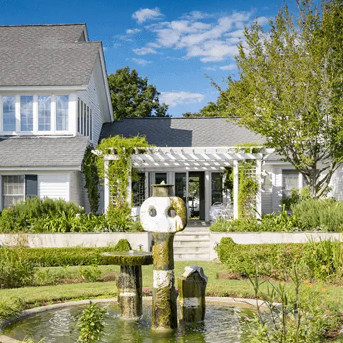 A two-story white house with large windows, a gray roof, and a white pergola over a patio. In the foreground, there's a stone fountain surrounded by lush green gardens and manicured shrubs under a blue sky.
