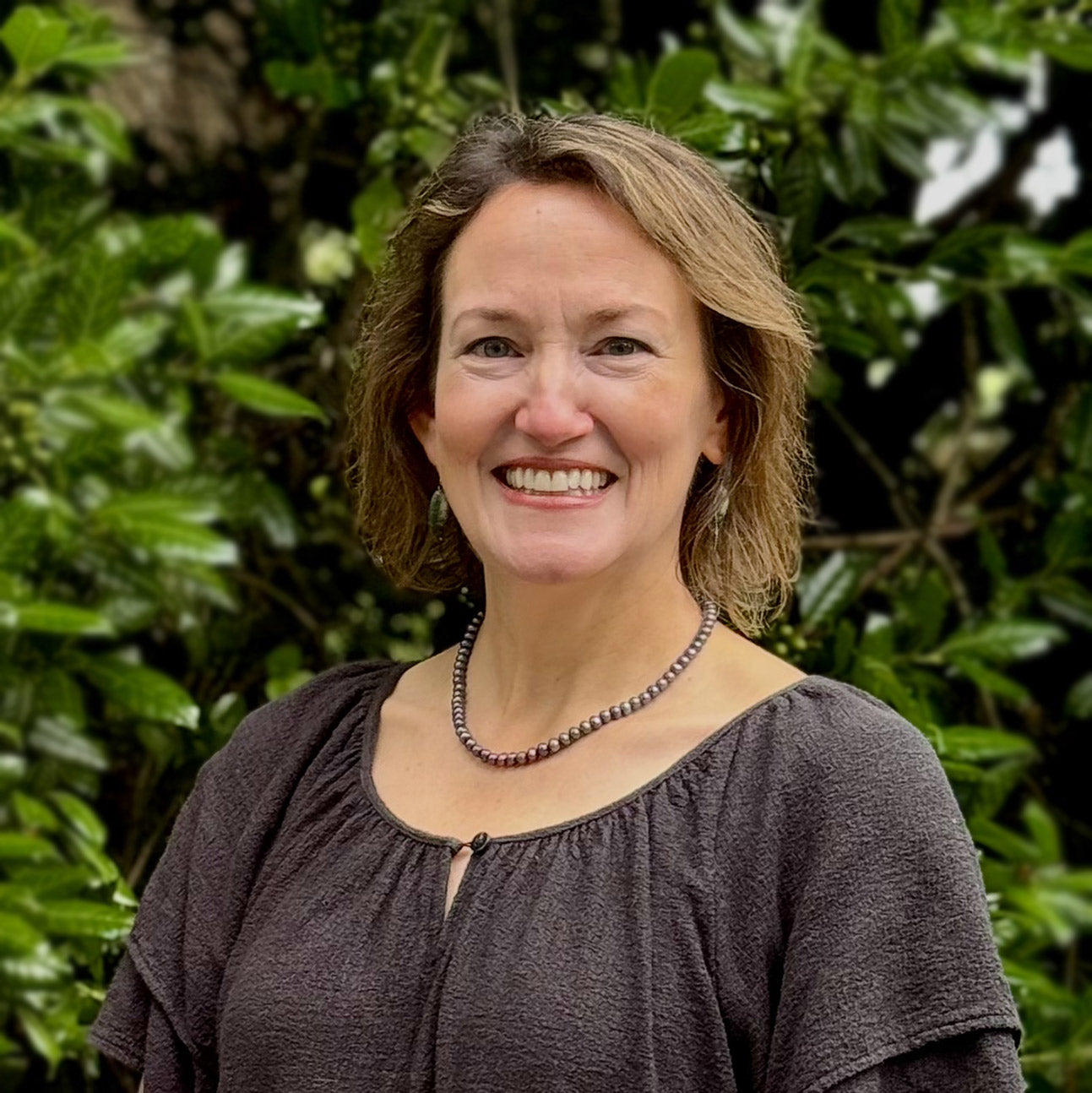 A woman with wavy, shoulder-length hair, wearing a necklace and a textured dark blouse, smiles while standing outdoors in front of leafy green plants.
