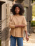 A person with curly hair stands outside in front of a building, wearing FRANK & EILEEN's Effie Long Sleeve Funnel Neck Capelet paired with blue jeans. They have a relaxed pose, accentuated by the soft drape of the capelet. The background features a wooden structure and lush greenery.