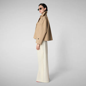 The SAVE THE DUCK - GRETA TRENCH JACKET is a tan trench coat with a wrap-style silhouette, featuring a wide collar, asymmetric flap on the right side, and long sleeves with buttoned cuffs. Its minimalist design stands out against a plain gray background.