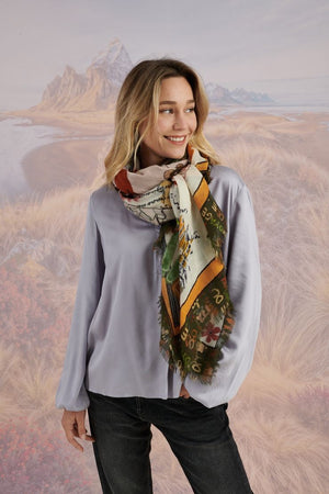 A woman with wavy blonde hair smiles while looking down. She wears a loose, light lavender blouse, dark pants, and the STORIATIPIC EUGENIE WOOL SCARF. Behind her is a scenic landscape of mountains, grass, and water.