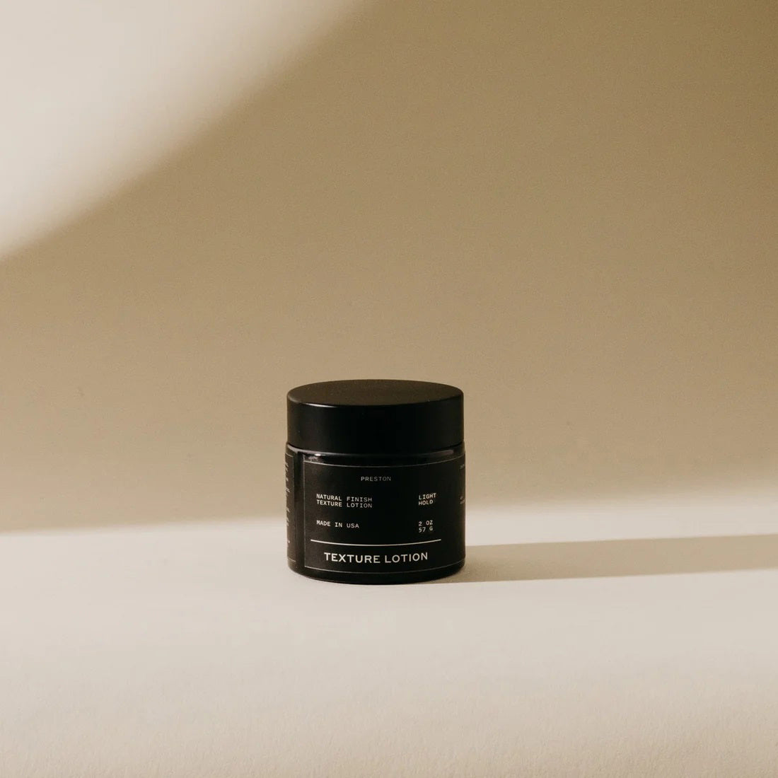 A close-up view of an open jar of PRESTON - TEXTURED LOTION reveals a thick, white cream with a matte finish. The jar has a black rim and a slightly reflective surface that contrasts with the smooth and glossy texture of the lotion inside.