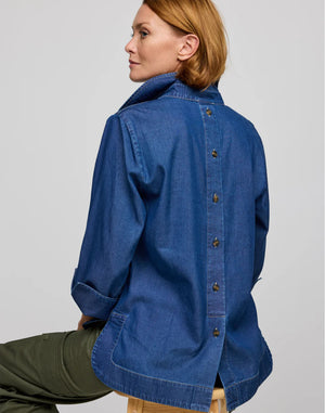 A person with short hair sits on a stool, facing away from the camera. They wear the breathable HINSON WU AILEEN 3/4 sleeve top in denim, featuring buttons down the back, paired with green pants.