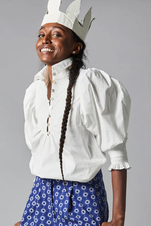 Rear view of a person wearing the Hope for Flowers Smocked Shirt, a bright red blouse with puffed sleeves, paired with blue jeans featuring pom-poms. Made from GOTS Certified organic cotton, this outfit embodies sustainable fashion against a gray background.