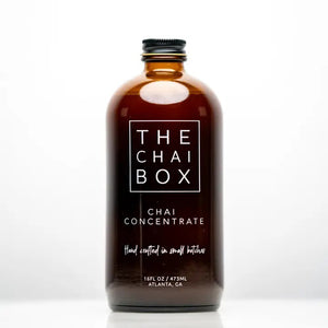 The Chai Box's brown glass bottle, prominently labeled with "THE CHAI BOX - CHAI CONCENTRATE," features text that reads "Hand crafted in small batches" and specifies a volume of 16 fl oz / 473 ml. Made with the finest spices, the label proudly displays "Atlanta, GA" at the bottom. The bottle is topped with a black screw cap.