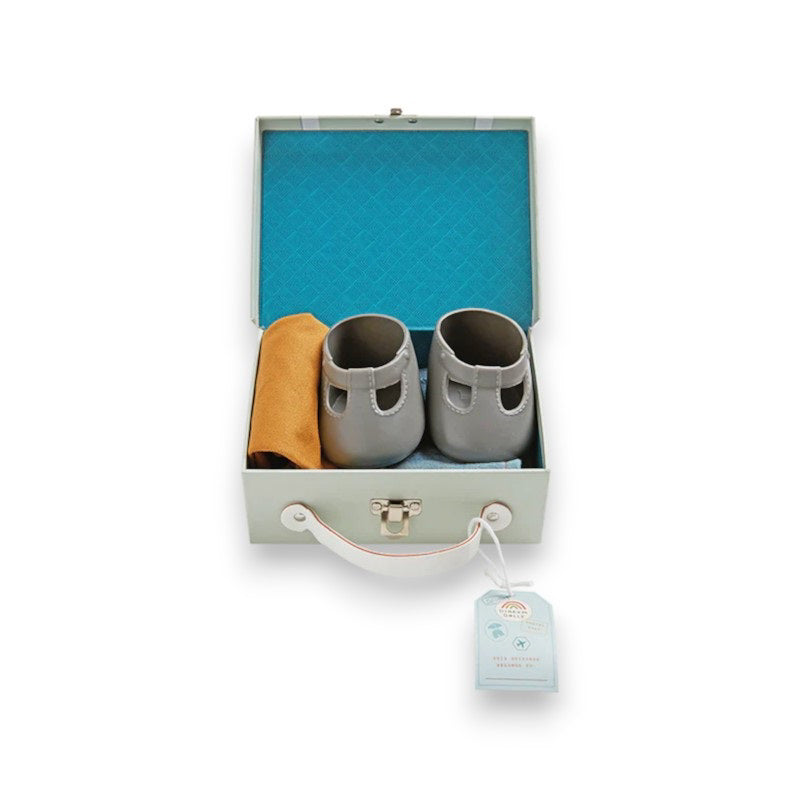 A Dinkum Doll from Olli Ella USA&#39;s Travel Togs collection, featuring dark hair and dressed in a denim jacket, mustard pants, and black shoes, stands on a wooden surface. Beside the doll is a teal See-Ya Suitcase with white trim and hardware. In the background, another larger teal suitcase is partially visible.