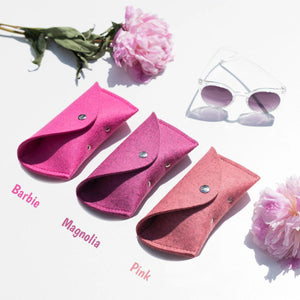 Three WOOLBERRY WOOL FELT GLASSES CASES in navy, bright, and light blue with magnetic closures are arranged on a white surface near clear sunglasses, framed by white-blue hydrangeas.