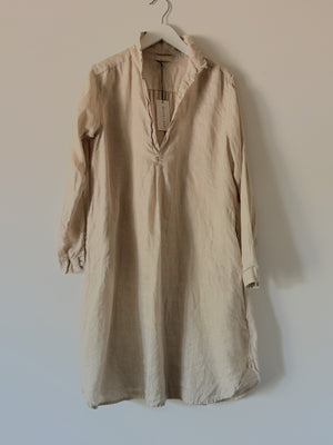 The CP SHADES - LARA LINEN DRESS in white, with long sleeves, hangs casually on a wooden hanger against a plain gray background. Made from 100% linen, this dress shirt features a deep V-neckline with a single button closure and possesses a relaxed, slightly wrinkled fabric. The sleeves are nonchalantly rolled up to the elbows, reflecting an essence of eco-friendly fashion synonymous with the CP SHADES brand.