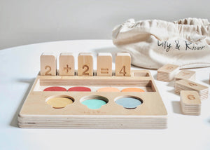 The "LITTLE NUMBERS GAME SET" by LILY & RIVER is a wooden educational toy influenced by Montessori principles. It features number blocks and colored disks on a white surface. The blocks display a simple math equation (2 + 2 = 4), functioning like a children's calculator. In the background, there is a light-colored fabric bag with the text "Lily & River" partially visible.
