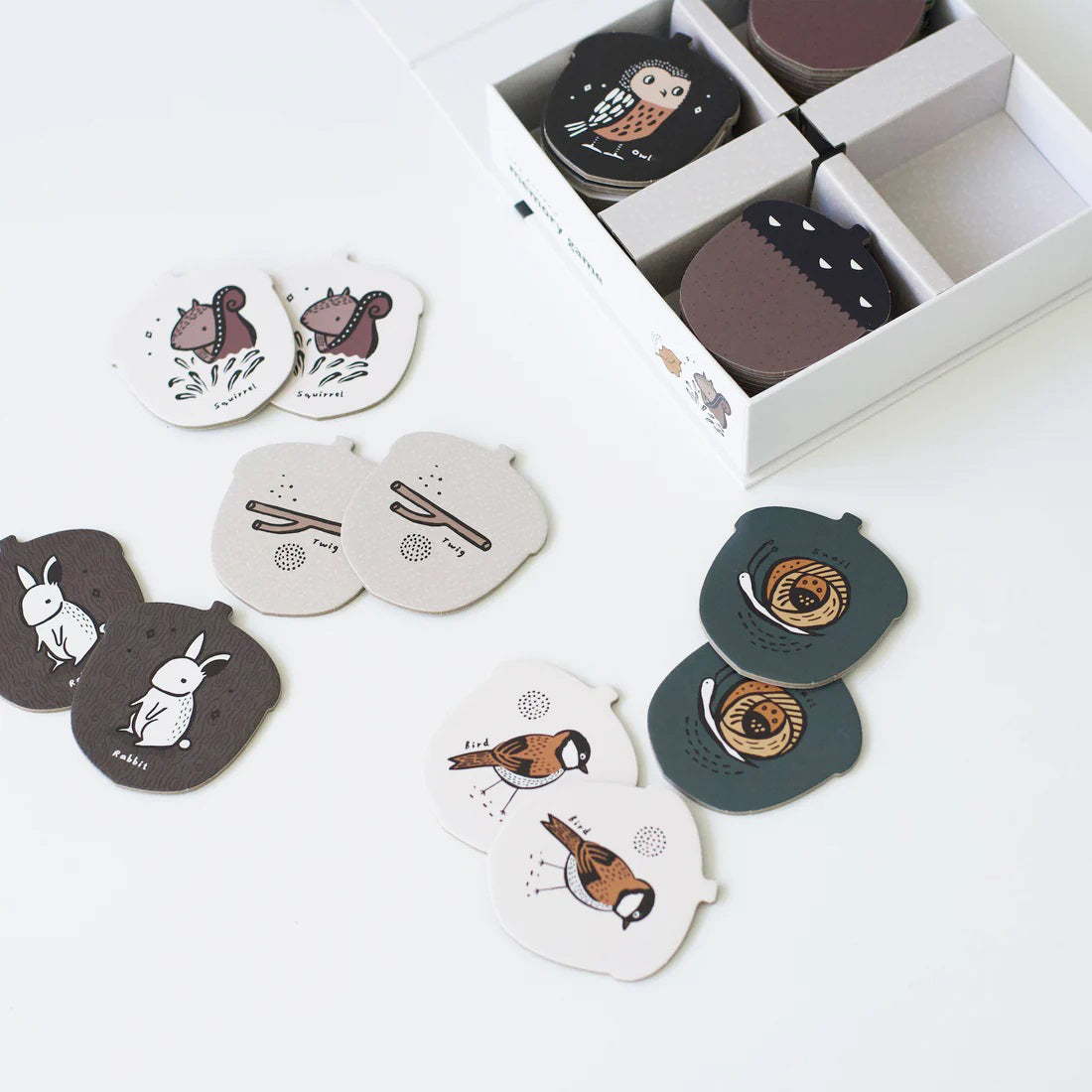 The WEE GALLERY - WOODLAND MEMORY GAME from WEE GALLERY is laid out on a white surface, showcasing pairs of cards with illustrations of birds, owls, snails, and rabbits. An open box also holds some additional cards, making it perfect for boosting cognitive skills.