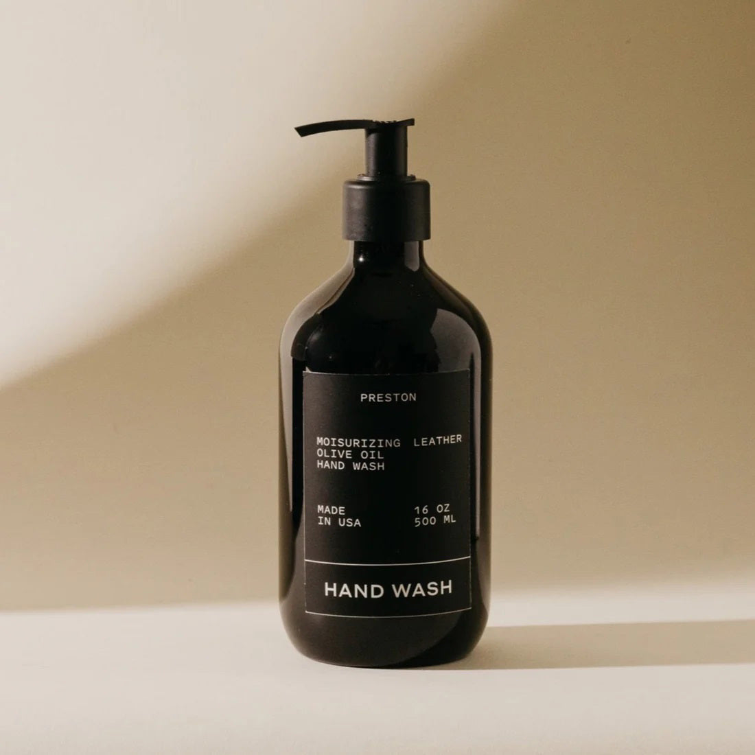 A black pump bottle with a label that reads &quot;PRESTON - HAND WASH 16OZ Moisturizing Olive Oil Hand Wash Amber, Sulfate-Free, Made in USA.&quot; The bottle is standing against a neutral background.