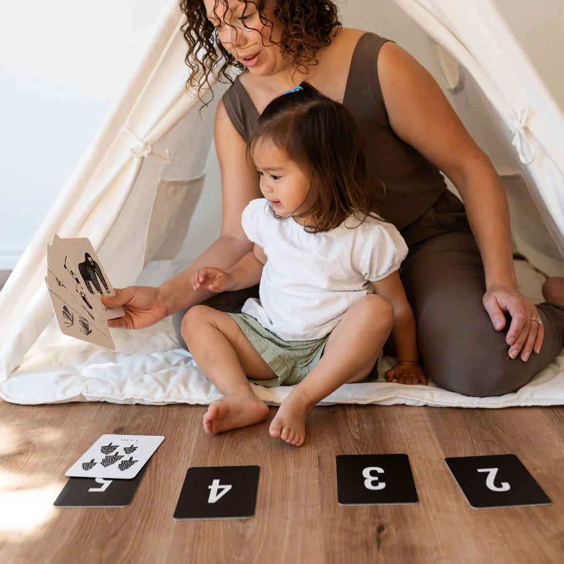 A woman and a young child sit together on the floor inside a white play tent. The woman is holding one of Wee Gallery&#39;s Nature Number Cards and showing it to the child. In front of them, four black cards with the numbers 1 to 4 from Wee Gallery&#39;s Nature Number Cards set are lined up on the floor.