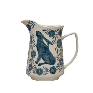 The HAND PAINTED STONEWARE PITCHER WITH RABBIT & FLOWERS from CREATIVE COOP features a charming blue design of a rabbit seated on its hind legs, surrounded by floral and leafy patterns. This 3-quart white and blue ceramic pitcher, complete with spout and handle, beautifully showcases the intricate Rabbit & Flowers decorations.