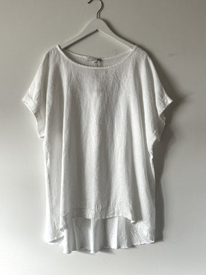 A short-sleeved, white linen-cotton top hangs on a white hanger against a light-colored wall. Made in the USA, the CUT LOOSE - ONE SIZE SEQUIN TRIM TOP boasts a loose fit and a slightly longer hem at the back.