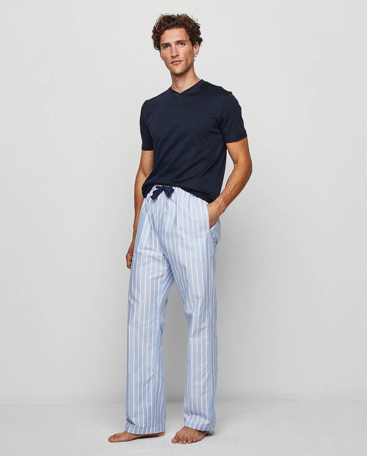 A man stands barefoot against a plain background, wearing the MIRTO 1956 - OXFORD PAJAMA SET by MIRTO 1956, featuring a dark blue top and light blue and white striped cotton pants with a comfortable drawstring waist.