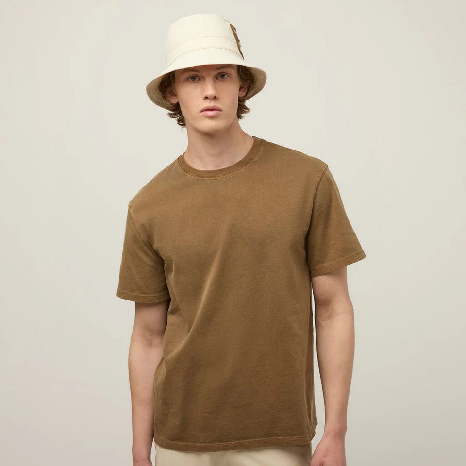 A young man in a beige bucket hat and a TILLEY Mid Weight Sunwashed T-Shirt in Peat stands against a light background, looking at the camera with a neutral expression.