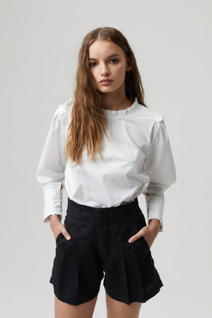 A young woman with long, wavy brown hair stands against a plain gray background. She is wearing the LANHTROPY - JANE COTTON POPLIN SHIRT from LANHTROPY, a white long-sleeved blouse made of 100% cotton poplin, featuring ruffled details at the neck and cuffs, paired with black high-waisted shorts. Her hands are in her pockets, and she has a neutral expression.