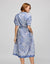 A person with shoulder-length hair wears the HINSON WU Luca Elbow Sleeve Lace Print Dress, a blue and white breathable dress featuring short sleeves, a belted waist, and a hand in a pocket against a plain background.