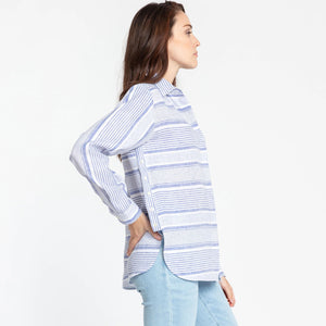 A person with long brown hair is wearing the HINSON WU - GEMMA LONG SLEEVE LINEN SHIRT, which features a light pink and white striped pattern and functional side button details. They have paired it with light blue jeans and are posing against a plain white background, looking directly at the camera with a neutral expression.