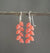 A pair of ANN LIGHTFOOT - CAMELIAN LEAF PETAL EARRINGS featuring sterling silver earwires with clusters of small, leaf petal-shaped beads in a vibrant coral color. The earrings are hanging on a dark, horizontal rod against a plain gray background.