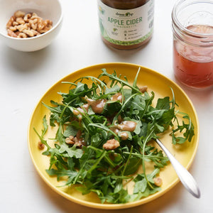 A 10 fl oz bottle of "Dress It Up Dressing - Apple Cider" by DRESS IT UP DRESSING is depicted. The label, featuring a green illustration of a woman in a dress, highlights its olive oil and simple ingredients. With the health benefits of apple cider vinegar, this vinaigrette sports a metallic cap against a white background.