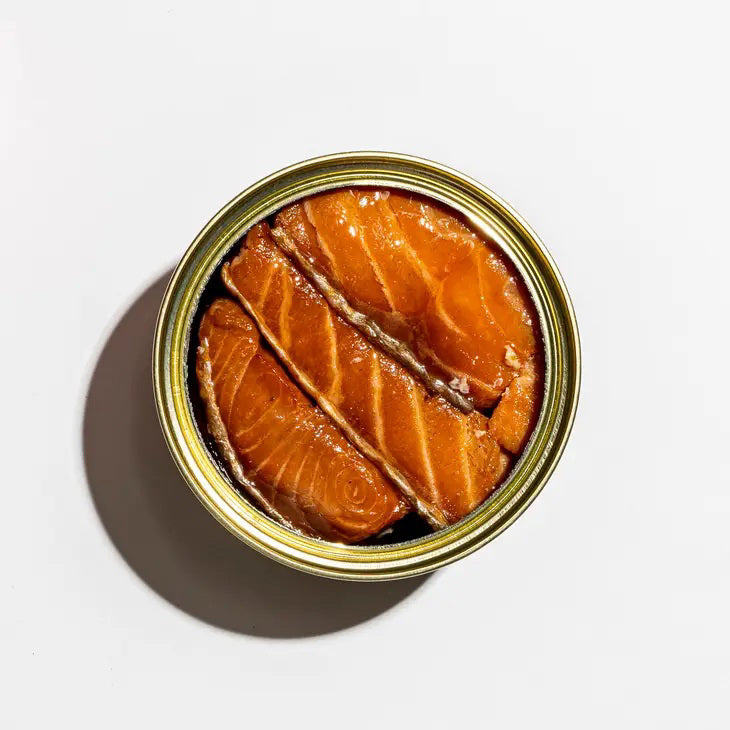 A red square box of FISHWIFE - SMOKED ATLANTIC SALMON is shown. The front showcases vibrant illustrations, including a woman holding a fish on a plate, decorative text, and labels such as "100% Traceable," "Responsibly Raised in America," emphasizing its origins from a family-run cannery that uses sustainable seafood.