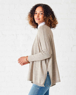 A person with curly hair is wearing the MERSEA - LET IT SNOW CATALINA CREWNECK SWEATER in beige over a white shirt and blue jeans, standing sideways against a white brick wall. They are smiling and looking at the camera.